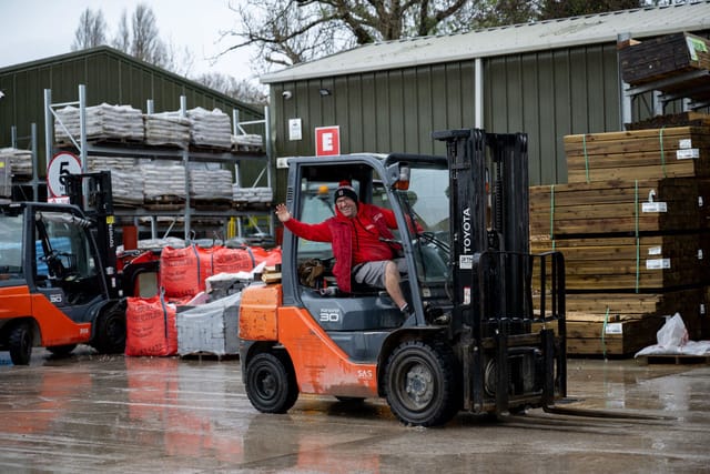 A worker in a red jacket driving a forklift at a warehouse loading zone.