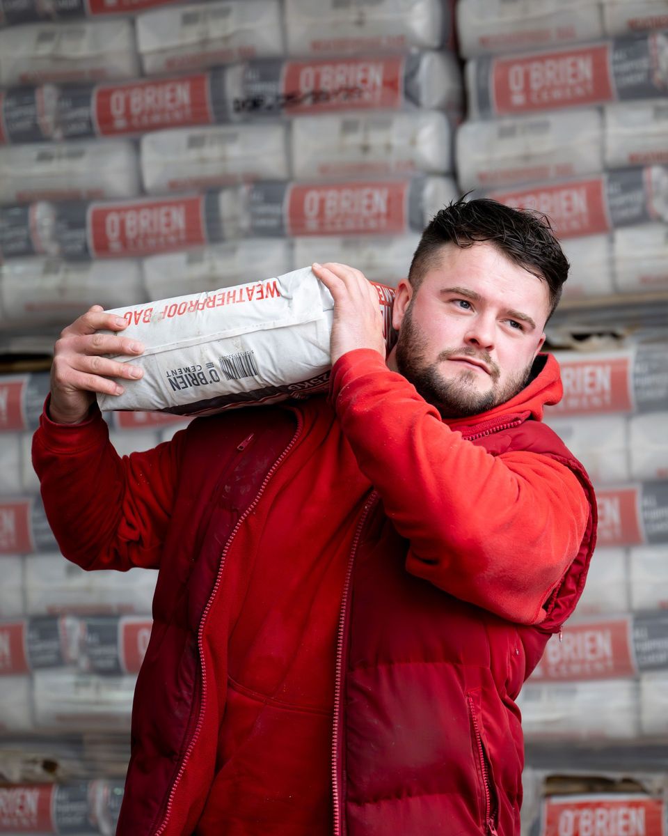 Man in red jacket carrying a bag of 'mastercrete' cement on his shoulder in a warehouse.