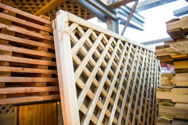 Stacks of wooden lattice panels and planks in a lumberyard, with a clear blue sky in the background.