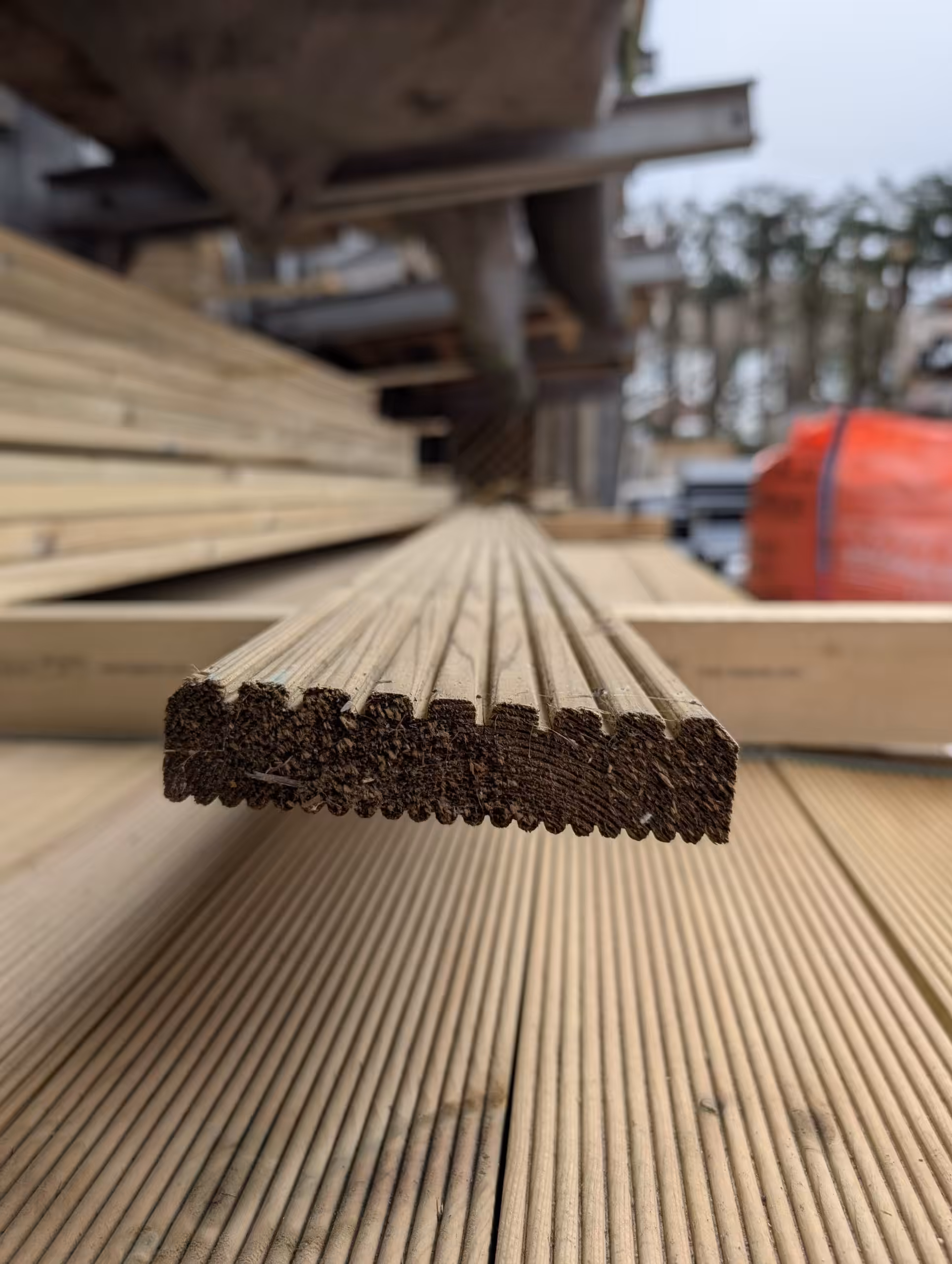 Treated Timber Decking Board - 00002 A close-up view of stacked Treated Decking Boards at a construction site, with additional building materials and trees visible in the background.