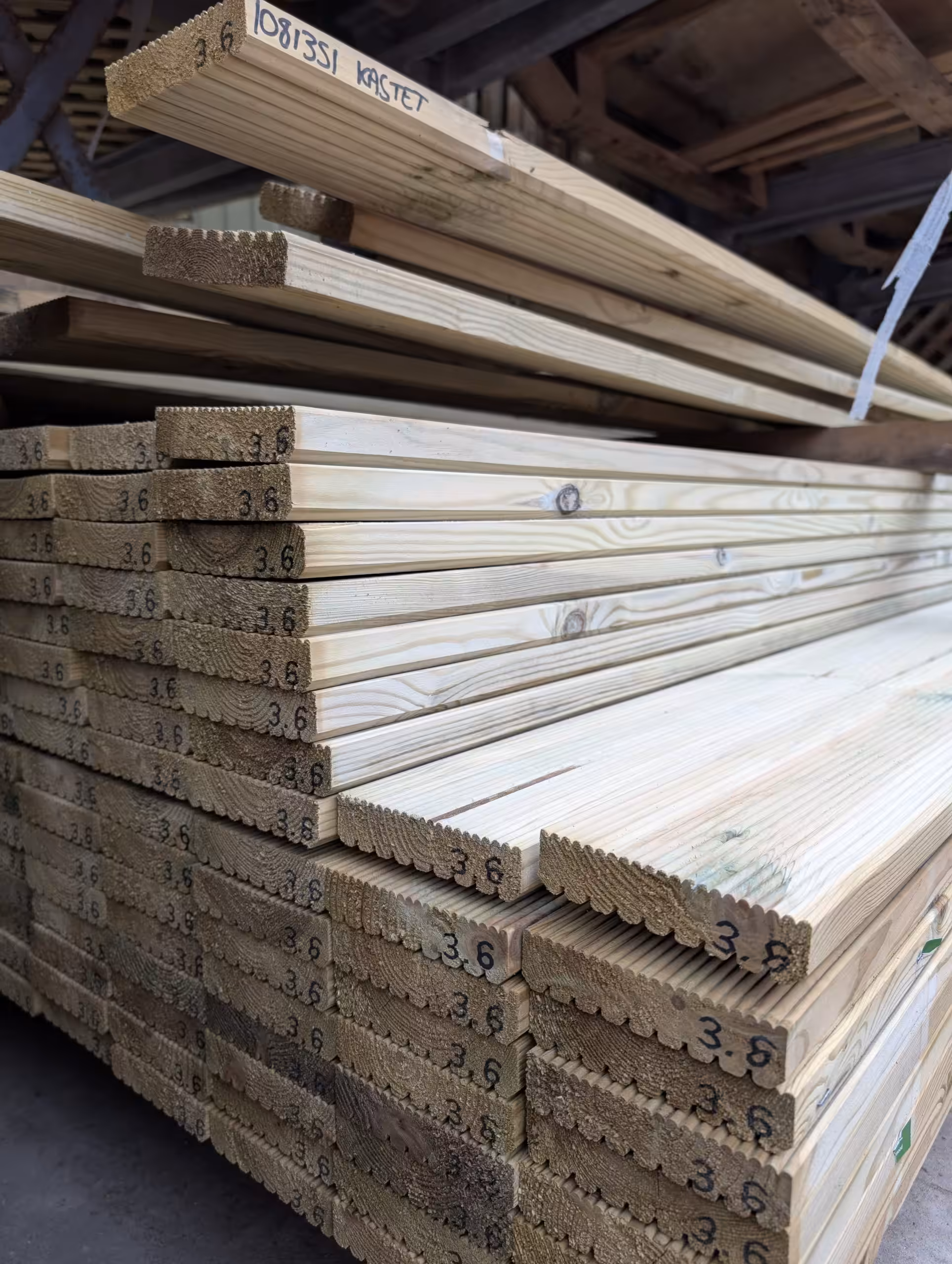 Treated Timber Decking Board - 00005 Treated Decking Boards with notched edges are arranged in rows with handwritten measurements on the ends and stored in an indoor lumber area.
