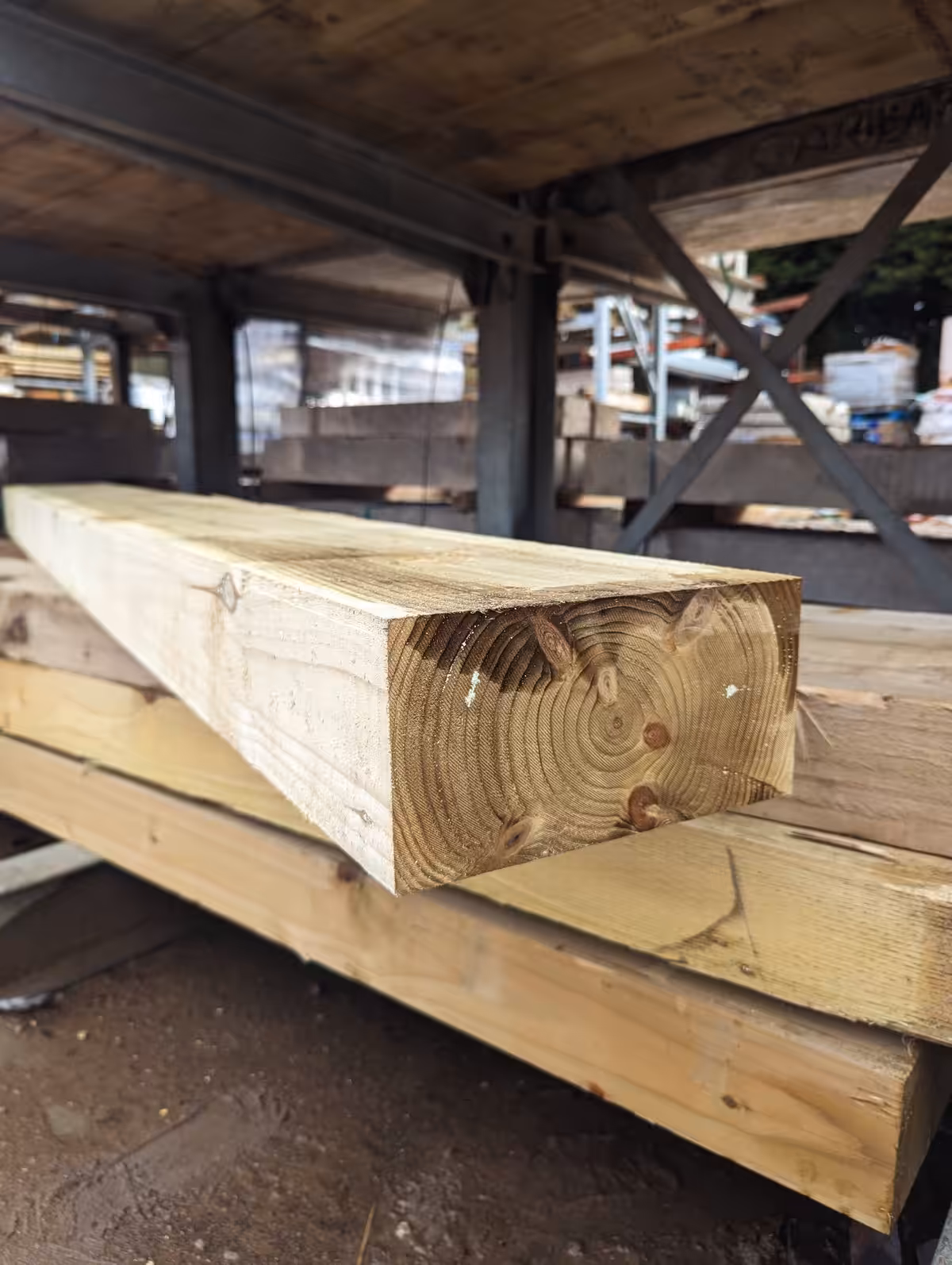 Stacked wooden beams with visible grain patterns stored on metal shelving in an outdoor lumber yard.