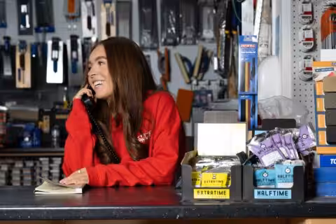A person in a red sweater is smiling and talking on a phone behind a counter in a hardware store, surrounded by various tools and products on display.