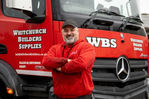 Person in a red hoodie and black cap stands smiling with arms crossed in front of a red Whitchurch Builders Supplies truck.