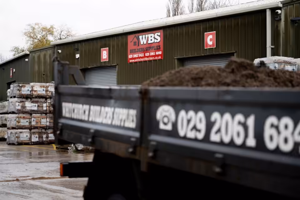A truck loaded with building materials is parked in front of a building labeled “WBS Builders Supplies” with signs marked B and C.