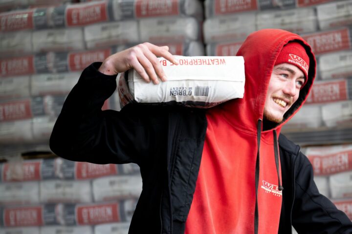 A man in a red hoodie and beanie smiles while carrying a bag of WeatherPro cement on his shoulder. Stacks of similar bags are in the background.