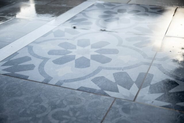 Close-up of wet, patterned tiles with a geometric floral design. Reflections and water create a glossy surface.