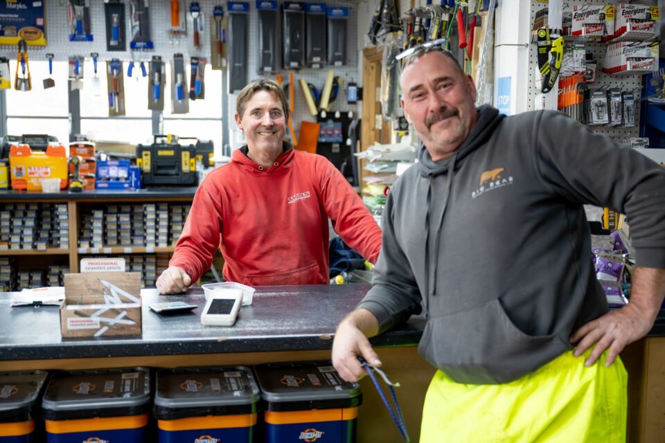 Two men smiling in a hardware store, standing behind a counter with tools and equipment displayed on shelves in the background.